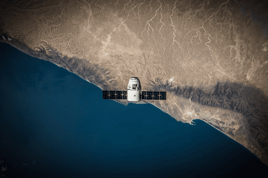 A spacecraft with extended solar panels above a rugged desert coastline and deep blue water